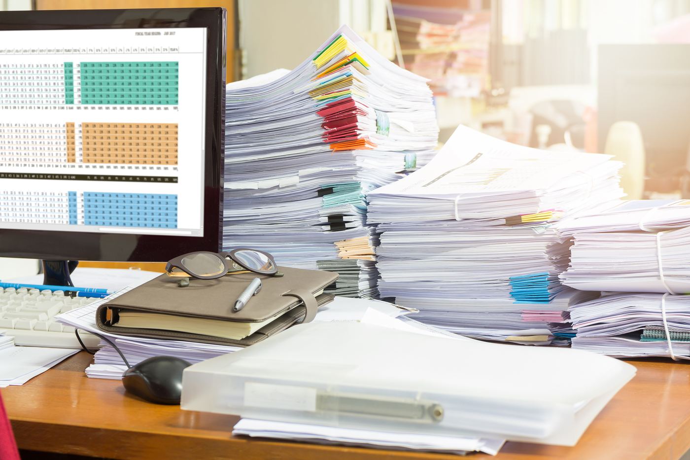 Office desk piled with paperwork next to a laptop showing a spreadsheet, representing manual data entry busywork