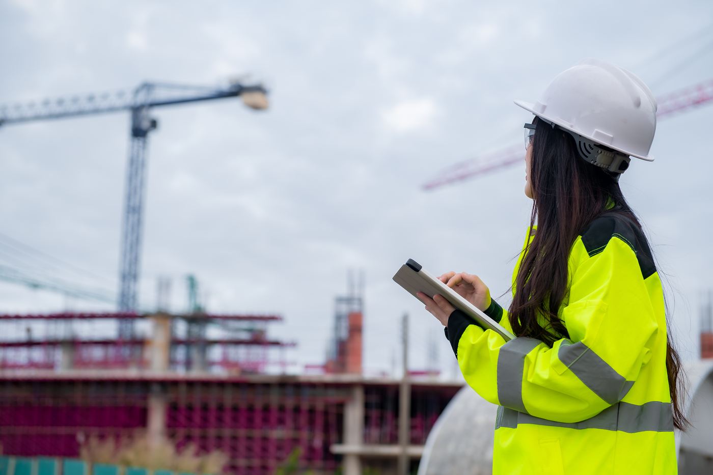 Construction engineer in a hard hat reviewing job-site data on a tablet with cranes in the background