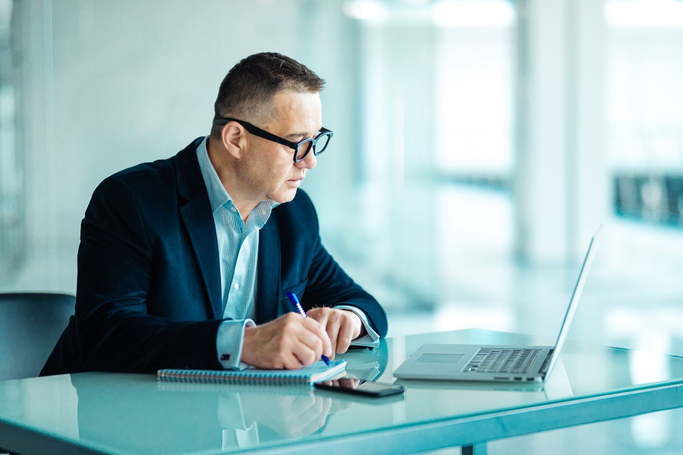 Consultant taking notes beside an open laptop in a modern glass-walled office